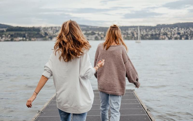 two women walk to the edge of a dock, in a body of water, looking toward the city on the other shore